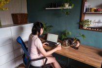 Vue latérale de documents de lecture indépendants asiatiques sur ordinateur portable assis à table pendant le travail à distance — Photo de stock