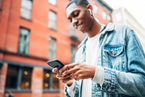 From below crop cheerful content African American guy in denim trendy jacket surfing modern mobile phone during city stroll — Stock Photo