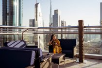 Giovane viaggiatrice che beve caffè e gode di una vista mozzafiato sulla città di Dubai con l'architettura contemporanea, mentre seduto sulla terrazza panoramica della caffetteria moderna — Foto stock