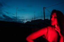 Délicieuse jeune hispanique ethnique femelle en lingerie regardant la caméra tout en se reposant sur la terrasse sous la lumière rouge néon la nuit avec ciel bleu foncé — Photo de stock