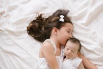 Visão superior da menina alegre abraçando bebê adorável enquanto deitado em cama macia em casa — Fotografia de Stock
