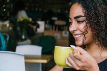 Zufriedene junge hispanische lockige Brünette trinkt aromatischen Kaffee aus gelbem Becher, während sie allein auf der Caféterrasse in der Nähe grüner Büsche chillt — Stockfoto