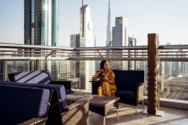 Young female traveler drinking coffee and enjoying breathtaking view of Dubai city with contemporary architecture while sitting on rooftop terrace of modern coffee shop — Stock Photo
