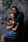 Zarter ethnischer Mann umarmt lächelnde Frau auf dunklem Hintergrund im Studio und blickt in die Kamera — Stockfoto