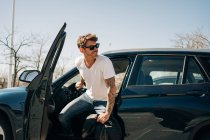 Fröhlicher Mann mit Sonnenbrille steigt an sonnigen Tagen aus modernem Auto und schaut weg — Stockfoto