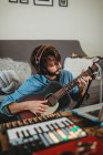 Young man in headphones playing on guitar near table with laptop and synthesizer at home looking at camera — Stock Photo