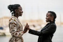 Damas afroamericanas sonrientes de moda que pasan tiempo juntas y se dan la mano en el parque en un día brillante - foto de stock