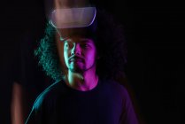 Восхитительный латиноамериканец с вьющимися волосами и современными очками виртуальной реальности, стоящий в студии на черном фоне с неоновыми огнями — стоковое фото
