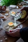 Cropped unrecognizable person preparing fresh cooked ramen noodles with tofu, eggs and vegetables with bottle of broth on a wooden table — Stock Photo