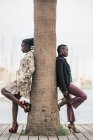 As senhoras americanas africanas na moda na moda que passam o tempo em conjunto e apoiam-se no tronco de uma palmeira no parque no dia brilhante — Fotografia de Stock
