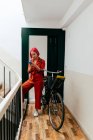 Jovem mulher elegante em terno vermelho com mochila usando smartphone enquanto está de pé com bicicleta na escada — Fotografia de Stock