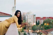 Молодая латиноамериканка в повседневной одежде опирается на перила и смотрит в камеру, расслабляясь на балконе вечером в городе — стоковое фото