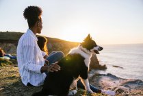 Vista lateral da jovem dona afro-americana com o cão Border Collie passando tempo juntos na praia perto do mar acenando ao pôr do sol contemplando vistas — Fotografia de Stock