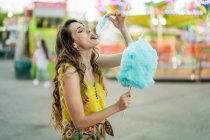 Vista lateral da fêmea infantil comendo doces de algodão azul doce enquanto se diverte e desfruta de fim de semana no parque de diversões no verão — Fotografia de Stock