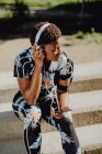 Молодая афроамериканка в разноцветной спортивной одежде слушает музыку в наушниках, сидя на каменной лестнице в парке — стоковое фото