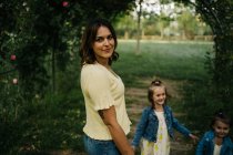 Young woman with adorable little daughters in similar outfits while walking together on green grassy lawn in summer park — Stock Photo
