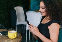 Side view of happy young Hispanic female browsing mobile phone while enjoying delicious cappuccino at table of street cafe — Stock Photo