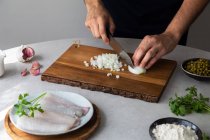 Unrecognizable man cook cutting onions with knife on a wooden board near peas and hake fillet with herbs during food preparation in kitchen — Stock Photo