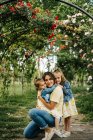 Feliz madre joven con lindas hijas con ropa similar disfrutando del soleado día de verano juntas mientras están sentadas en el parque - foto de stock