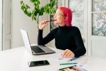 Ragionevole designer freelance donna in abito casual e occhiali che lavorano a tavola con computer portatile e disegno schizzi di moda con matite — Foto stock