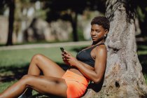 Africano mulher americana em sportswear segurando smartphone enquanto sentado na grama no dia ensolarado — Fotografia de Stock