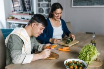 Mãe étnica alegre e filho adolescente com síndrome de Down sentado à mesa e cortando legumes enquanto prepara a salada para o almoço em casa — Fotografia de Stock