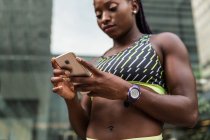 Cortar afro-americano fêmea em sportswear navegando smartphone moderno, enquanto de pé no fundo turvo da rua da cidade durante o treinamento ao ar livre — Fotografia de Stock