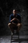 Delizioso elegante maschio etnico seduto su uno sgabello di legno in studio su sfondo scuro e guardando la fotocamera — Foto stock