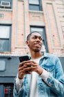 From below black positive young man in jeans outfit messaging on mobile phone while walking in city looking away — Stock Photo