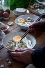 Обрізана невпізнавана людина готує свіжу варену локшину рамен з тофу, яйцями та овочами з паличками на дерев'яному столі — стокове фото