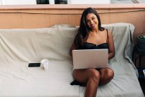 Улыбающаяся латиноамериканка с длинными волосами сидит на диване, смотрит в камеру и просматривает социальные сети на ноутбуке, отдыхая на балконе в выходные — стоковое фото
