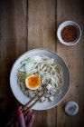 Von oben zugeschnittene unkenntliche Person, die frisch gekochte Ramen-Nudeln mit Tofu, Eier und Gemüse mit Essstäbchen auf einem Holztisch isst — Stockfoto