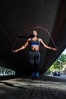 Afroamerikanerin in Sportkleidung hält Springseil in der Hand und blickt in die Kamera, während sie auf dem Bürgersteig der Stadtstraße steht — Stockfoto