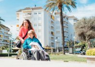 Доросла жінка штовхає інвалідне крісло з старшою матір'ю під час прогулянки по вулиці міста влітку — стокове фото