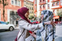 Seitenansicht von zufriedenen muslimischen Freundinnen in Hijabs, die auf der Straße stehen und sich umarmen, während sie einander anschauen — Stockfoto