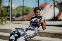 Молодая афроамериканка в разноцветной спортивной одежде слушает музыку в наушниках, сидя на каменной лестнице в парке — стоковое фото