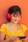 Contenuto internet per bambini navigando sul cellulare durante l'ascolto di canzoni da auricolari wireless su sfondo arancione — Foto stock