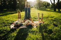 Mujer joven feliz y adorables hermanas pequeñas con vestidos similares tumbados en una manta sobre hierba verde mientras pasan el día de verano juntos en el parque y las piernas arriba - foto de stock