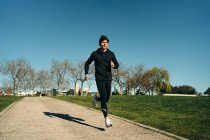 Взрослый бегун в спортивной одежде бегает по тротуару между газонами и смотрит вперед во время тренировки в городе — стоковое фото
