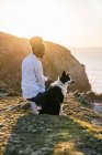 Vista laterale della giovane proprietaria afroamericana con il cane Border Collie che trascorre del tempo insieme sulla spiaggia vicino al mare che ondeggia al tramonto contemplando viste — Foto stock