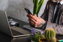 Crop unrecognizable tattooed male executive in checkered jacket text messaging on cellphone against laptop during telework — Stock Photo