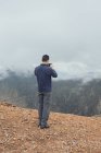 Обратный вид на неузнаваемого туриста-мужчину, стоящего на холме и фотографирующего удивительные высокогорья на смартфоне в туманный день во время отпуска в Уэльсе — стоковое фото