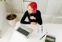 Von oben eine nachdenkliche freiberufliche Designerin in lässigem Outfit und Brille, die am Tisch mit Laptop arbeitet und Modeskizzen mit Bleistiften zeichnet — Stockfoto