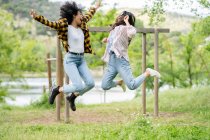 Couple of lesbian multiethnic females jumping above ground in woods and enjoying freedom together — Stock Photo