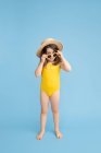 Corps complet de mignonne petite fille heureuse portant un maillot de bain jaune et un chapeau de paille avec des lunettes de soleil élégantes debout sur fond bleu et regardant la caméra — Photo de stock