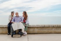 Счастливая семья веселится с пожилым отцом, сидящим в инвалидной коляске на набережной у моря в солнечный день — стоковое фото