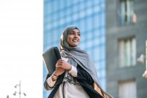 Empreendedora muçulmana alegre no hijab e com pasta de pé olhando para longe na rua — Fotografia de Stock