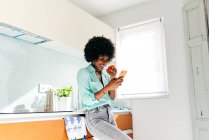 Desde abajo joven mujer afroamericana en ropa casual comer manzana y navegar por Internet en el teléfono móvil mientras está de pie en la cocina casera - foto de stock