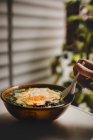Mano de persona anónima usando cuchara para tomar deliciosa sopa asiática con fideos y huevo frito en la terraza - foto de stock