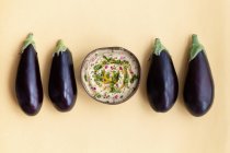 Vista dall'alto di melanzane fresche mature poste sul tavolo beige con ciotola di appetitoso piatto tradizionale Baba ghanoush — Foto stock